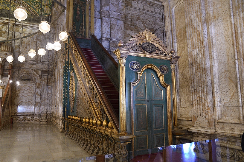 92 MAK_6975.jpg - Il minbar (pulpito rialzato) in legno finemente intagliato e decorato con dettagli dorati, caratterizzato da una scala con tappeto rosso e da una struttura d'ingresso con porte verdi.